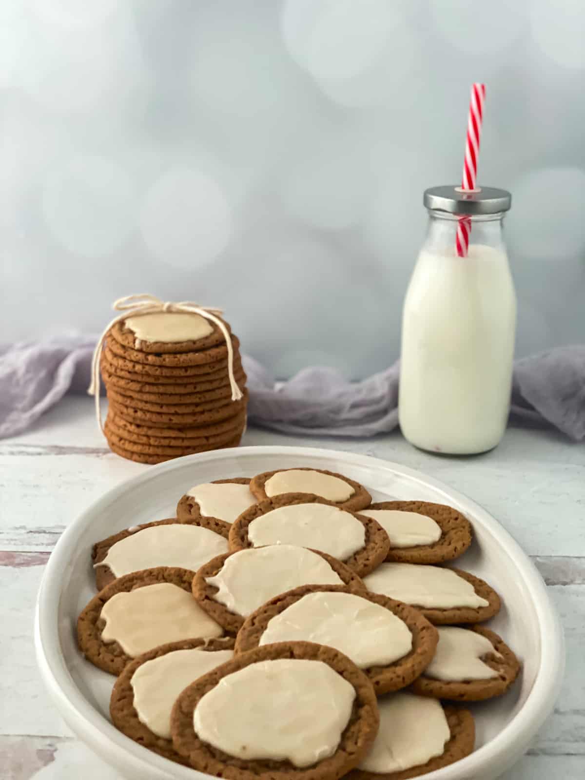 Iced cookies on a white plate with a stack of cookies and a bottle of milk in background.