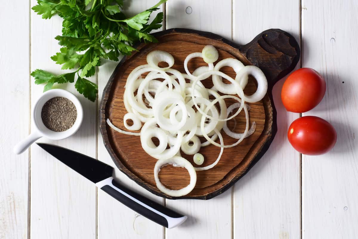 Sliced onions on a cutting board with tomatoes, parsley, and pepper.