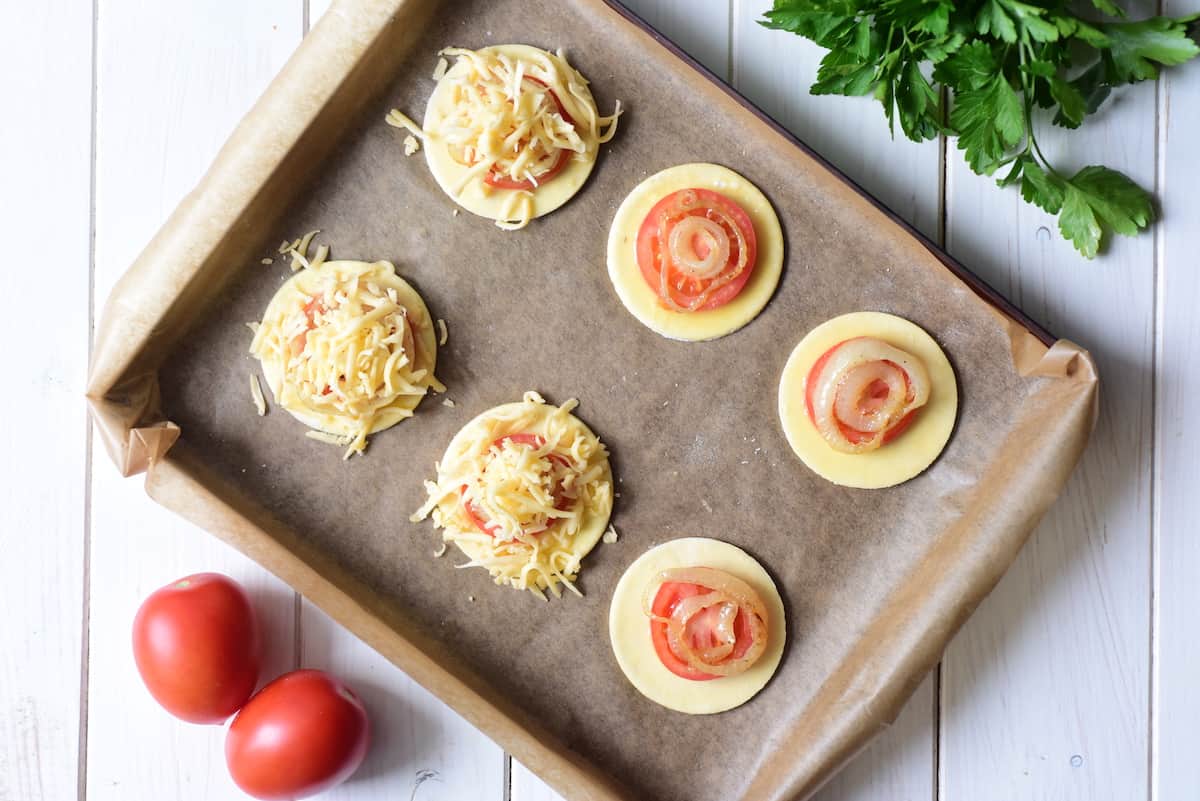 Making tomato tarts by adding tomato, onion, and cheese to pastry disc.