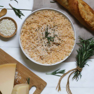 Risotto with cheese in a white bowl with rice, rosemary, bread, on a white table.