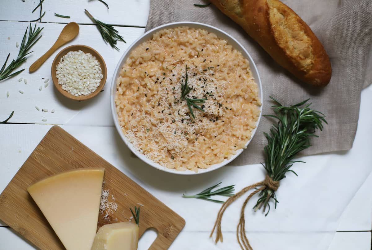 Arborio rice in a white bowl with bread, rosemary, rice, and cheese on white table.