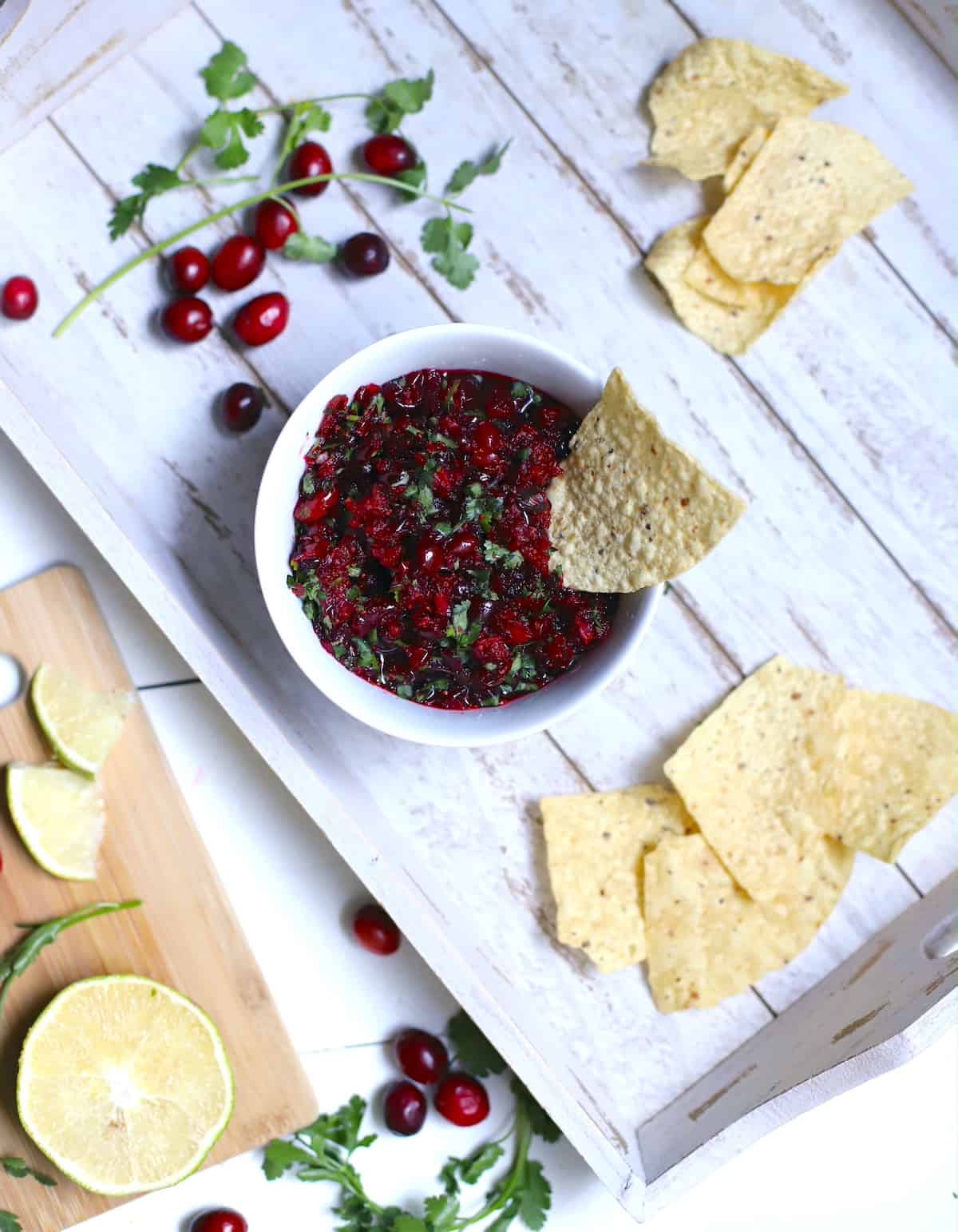 Cranberry salsa on a white try with cranberries, cilantro, limes, and chips.
