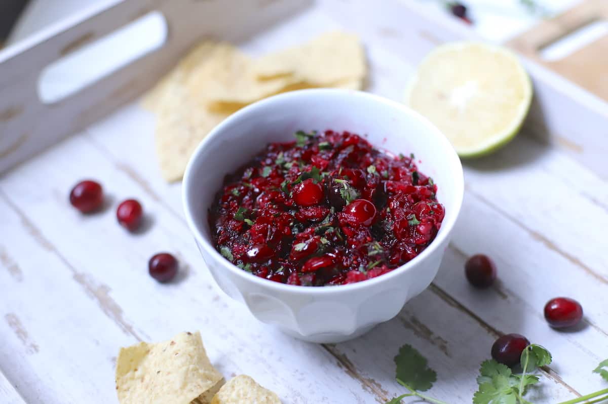 Cranberry salsa on a white tray with lime, chips, fresh cranberries, and cilantro.