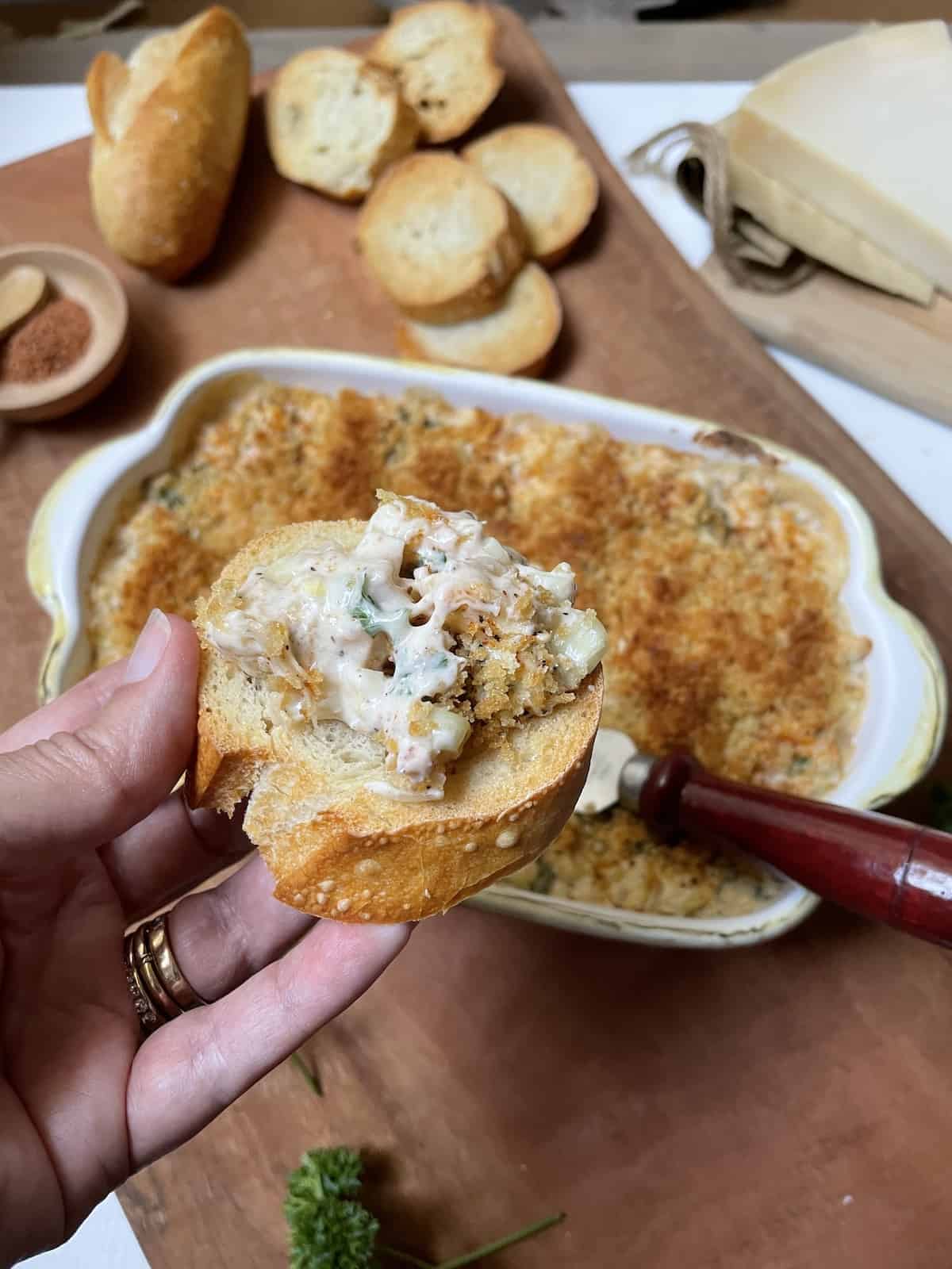 Shrimp dip on toast with more in a casserole dish on a wood board in background.