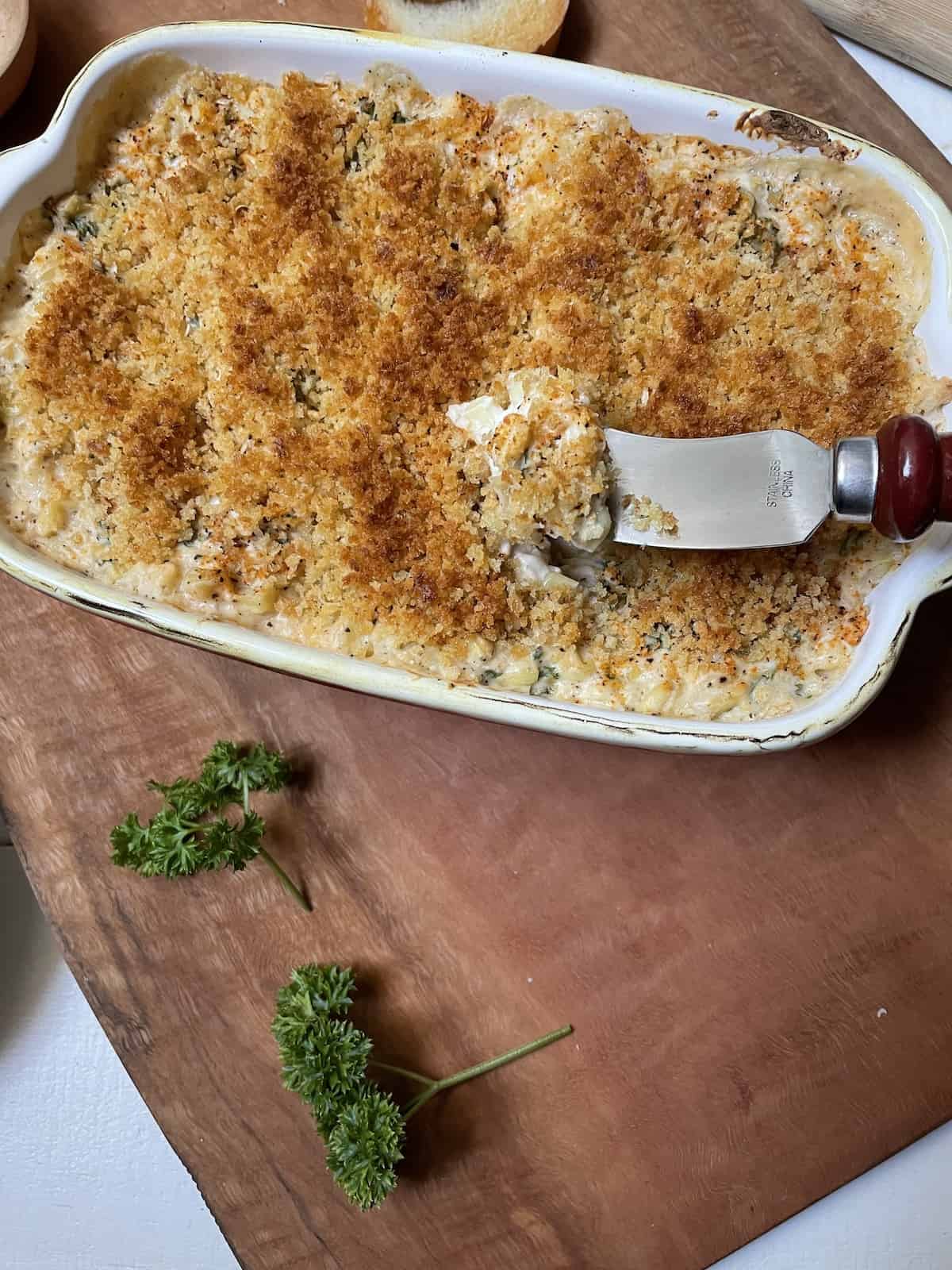 Shrimp dip in a casserole dish on a wood board with sliced bread, a spreader, parsley, and seasoning.
