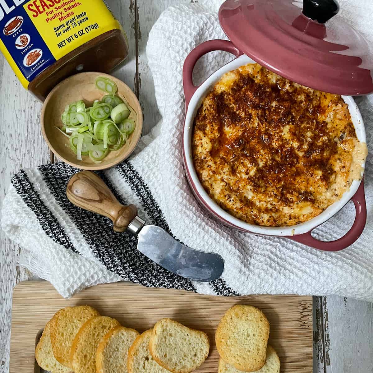 Crab dip in a dish on white towel with green onions and toasts.