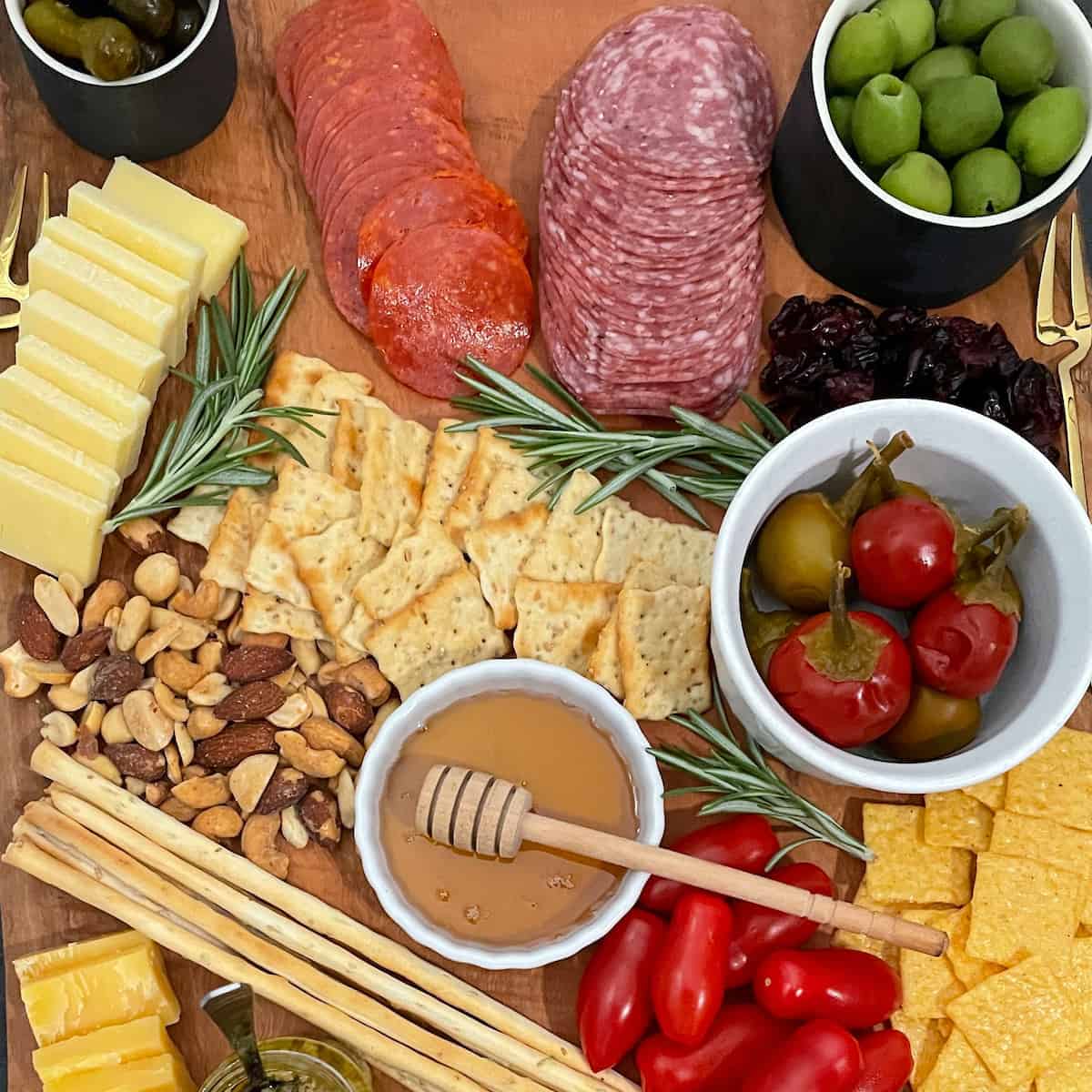 Charcuterie board with salame, pepperoni, cheeses, spreads, nuts, pickles, olives, and rosemary on a wood board.