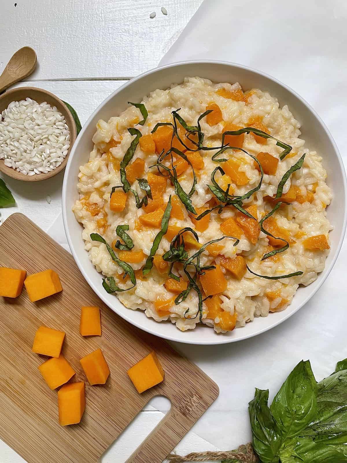 Butternut squash risotto in a white bowl on a white table beside a cutting board with butternut squash, a small bowl of uncooked rice,  and a bundle of fresh basil.