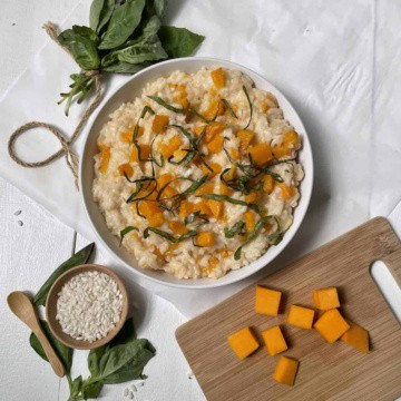 Butternut squash risotto in a white bowl on a white table beside a cutting board with butternut squash, a small bowl of uncooked rice, and a bundle of fresh basil.