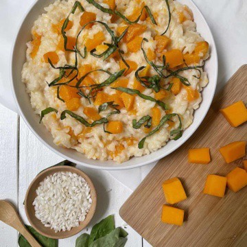 Butternut squash risotto in a white bowl on a white table with squash, small bowl of uncooked rice, and basil.