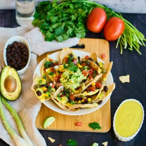 Nachos with guacamole, vegan cheese, tomato, cilantro, black beans, and spring onions on a wood board.