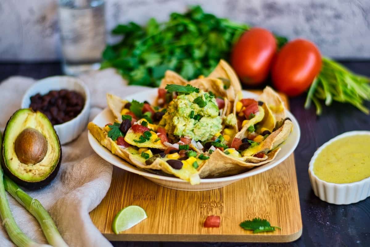 Nachos with guacamole, vegan cheese, tomato, cilantro, black beans, and spring onions on a wood board.