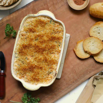Shrimp dip in a casserole dish on a wood board with sliced bread, a spreader, parsley, and seasoning.