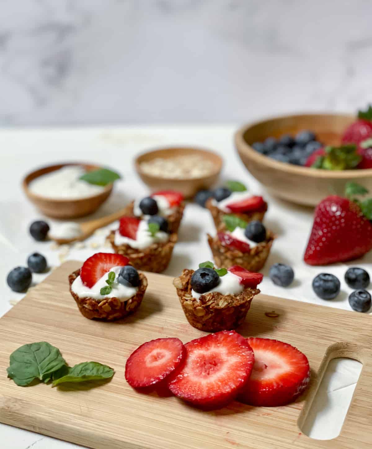 Oatmeal cups filled with yogurt and fruit on a cutting board on a white table with fruit and mint.