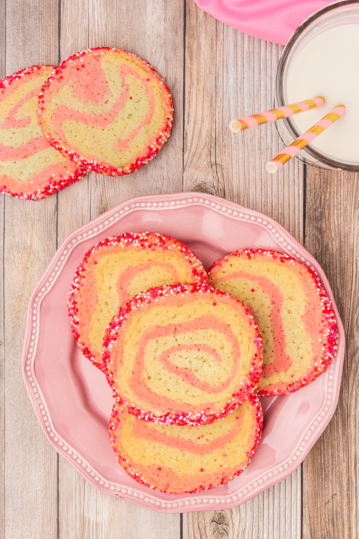 Cookies with a pinwheel swirl and sprinkles on outside on a pink plate with glass of milk.