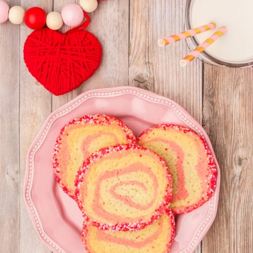 Cookies with a pinwheel swirl and sprinkles on outside on a pink plate with glass of milk.