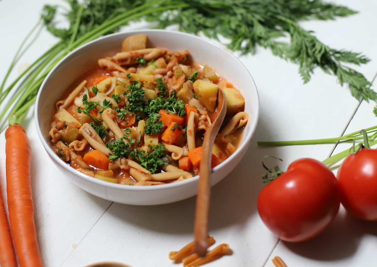 Minestrone in a white bowl with vegetables and pasta on white table.