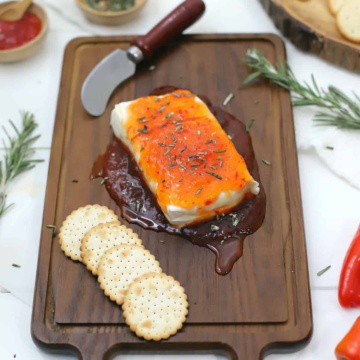 Cream cheese with red pepper jelly on a cutting board with red pepper jelly and rosemary in bowls and crackers on the side.