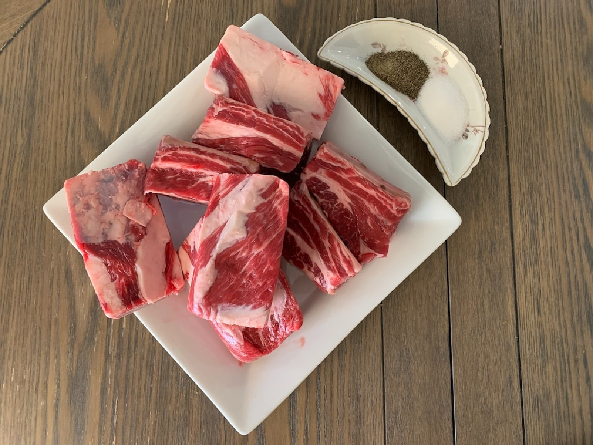 Raw short ribs on a white plate on a table with salt and pepper.