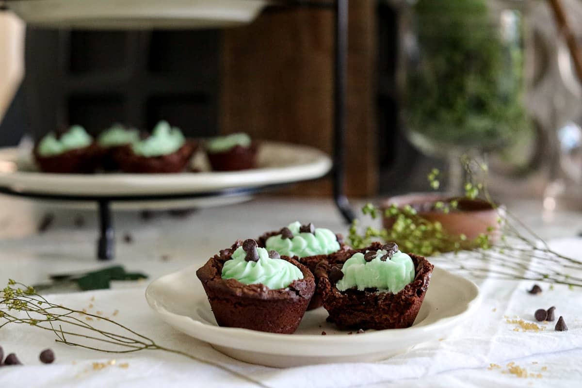 Three brownie bites with green buttercream filling on a white plate on a white tablecloth with more brownie bites in background.