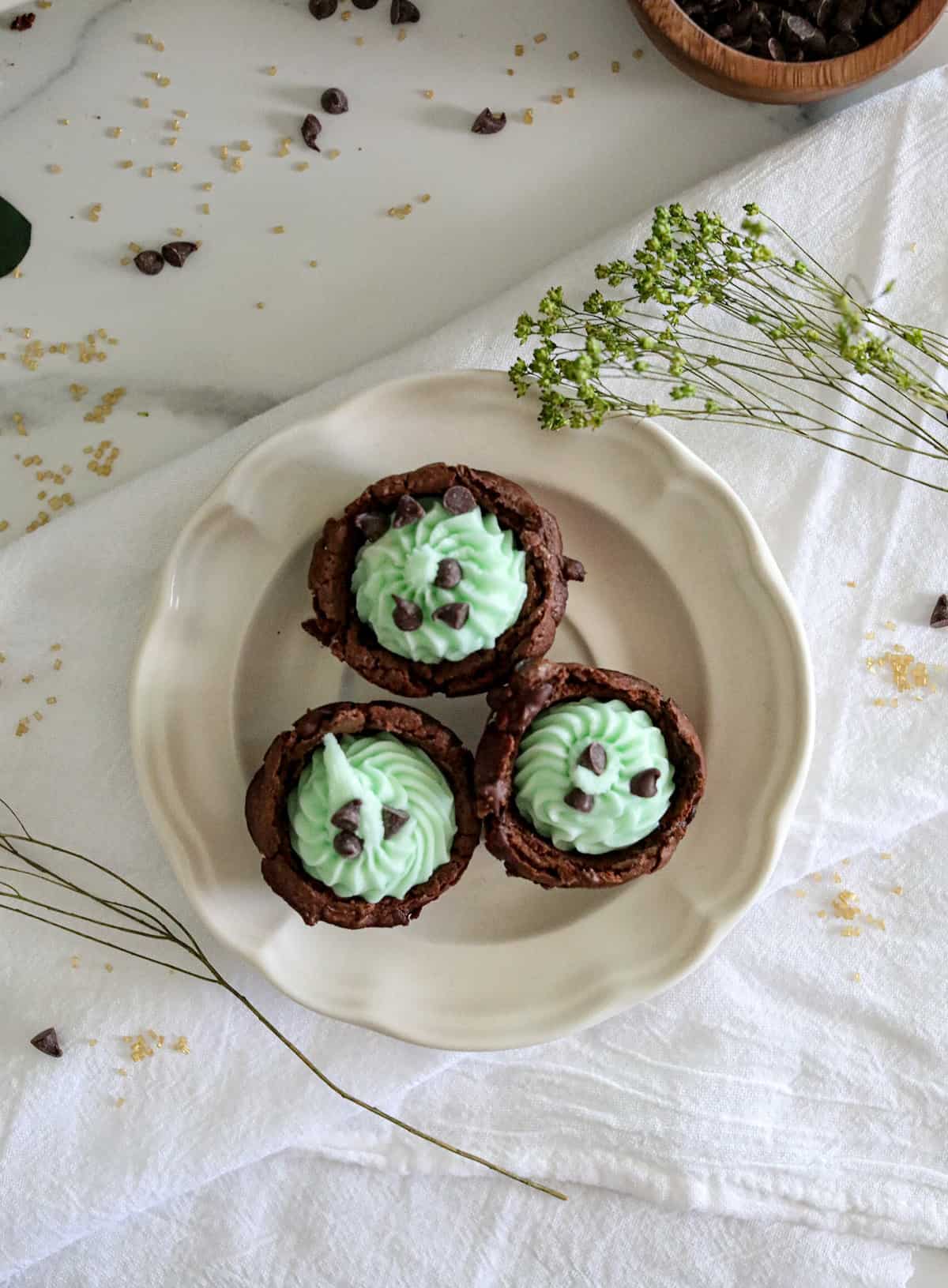 Three brownie bites with green buttercream filling on a white plate on a white tablecloth.