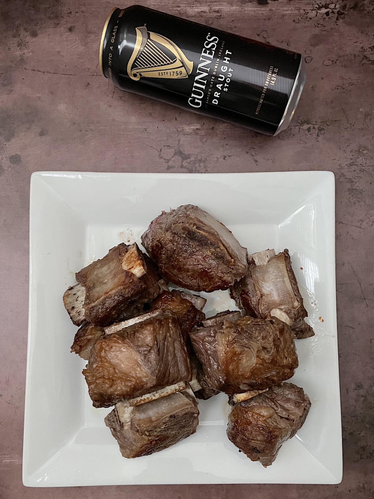 Seared short ribs on a white plate with a can of Guinness Stout.