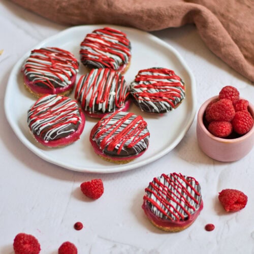 Raspberry sandwich cookies on a white plate on a white table with bowl of raspberries.