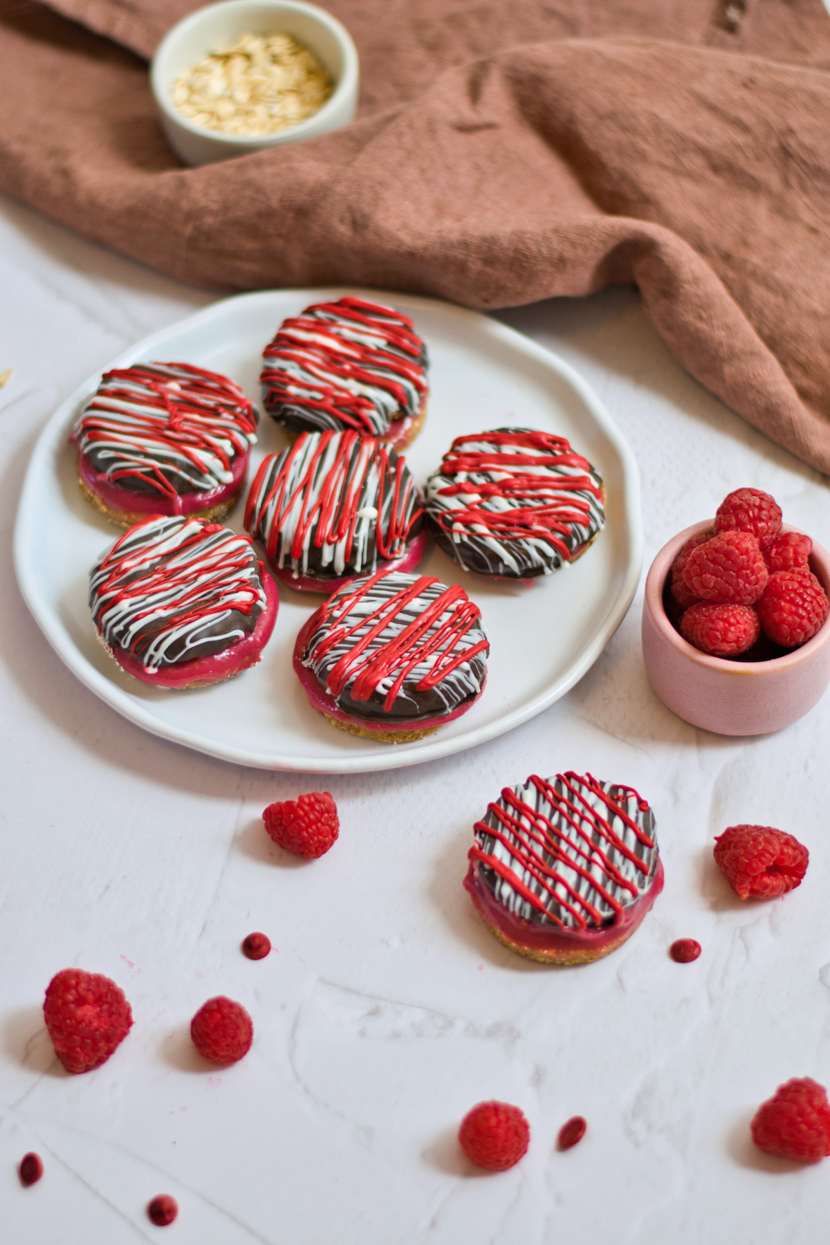 Raspberry sandwich cookies on a white plate on a white table with bowl of raspberries.