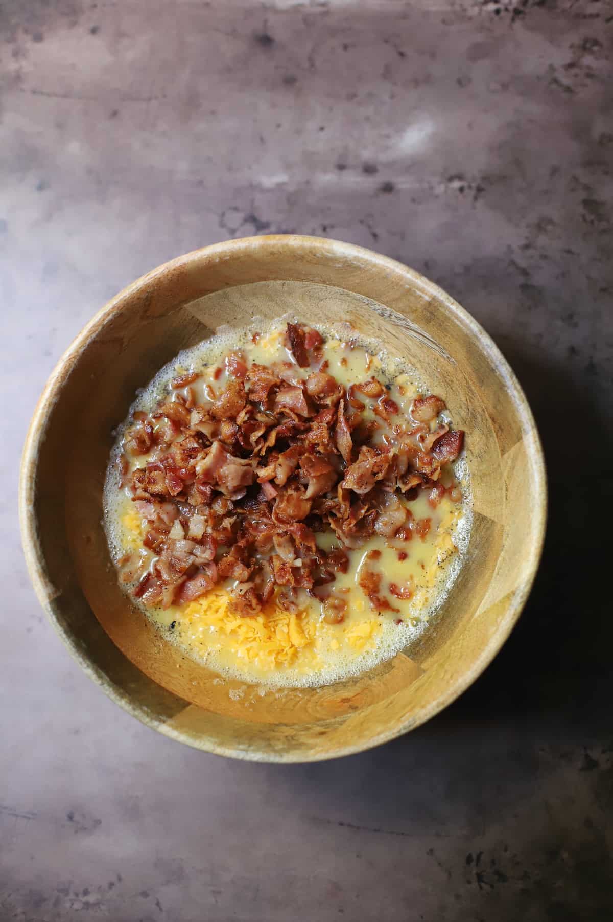 Eggs, hash browns, cheese, salt and pepper and bacon in a wood bowl.