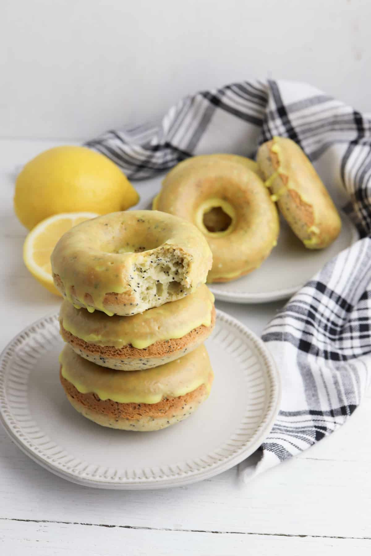 Lemon donuts stacked on a white plate with lemon and more donuts in background.