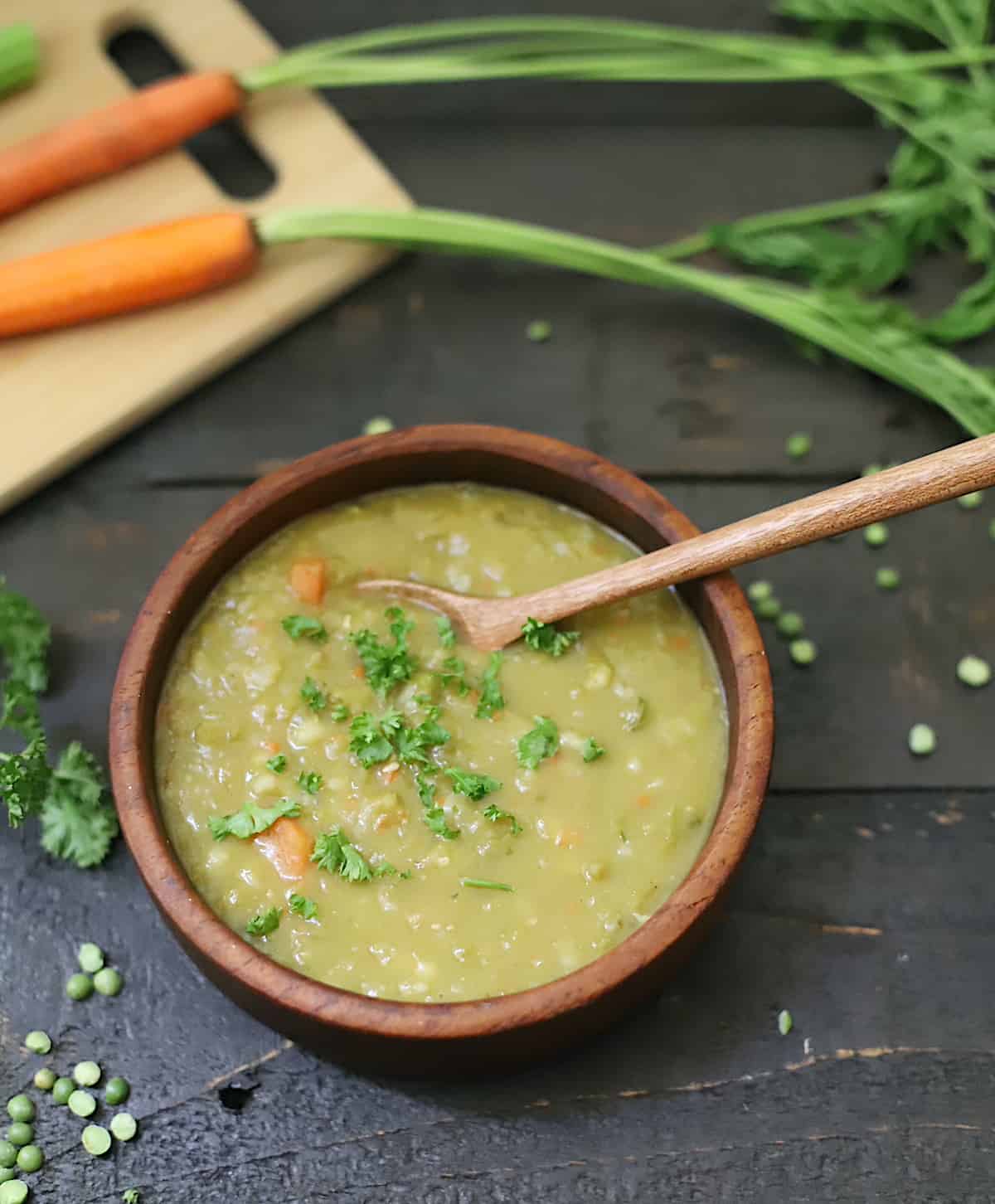 Bowl of pea soup on a black table with fresh carrots, celery, dried peas, and large pot to the side.