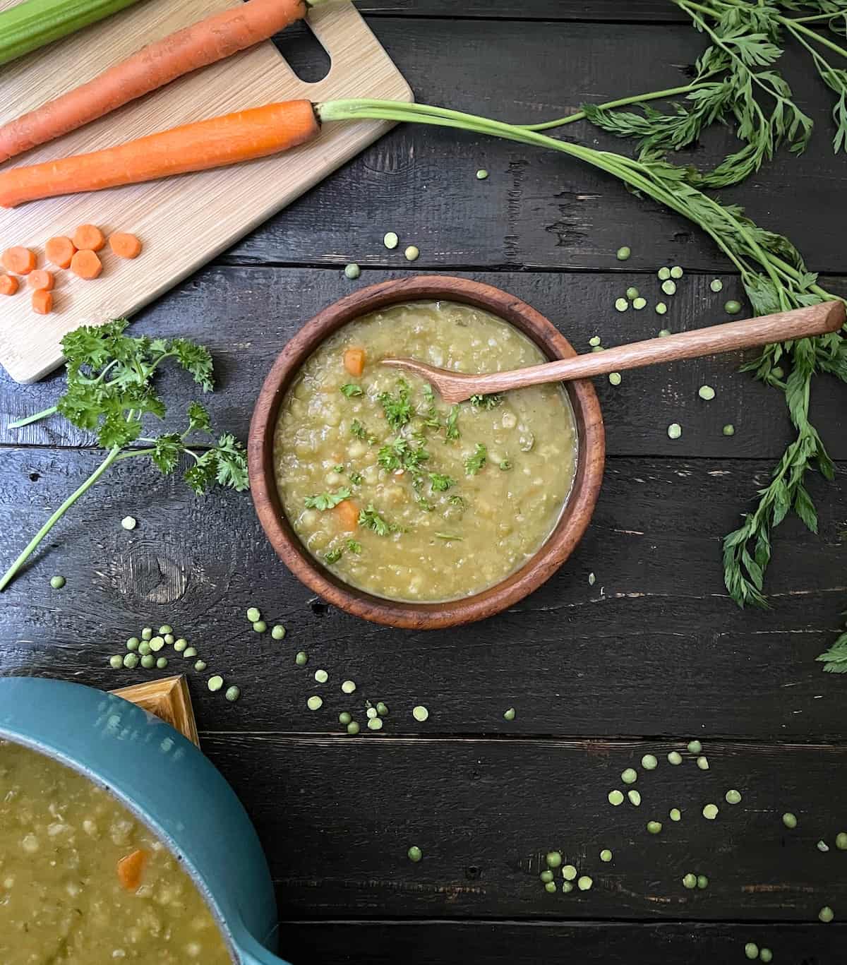 Bowl of pea soup on a black table with fresh carrots, celery, dried peas, and large pot to the side.
