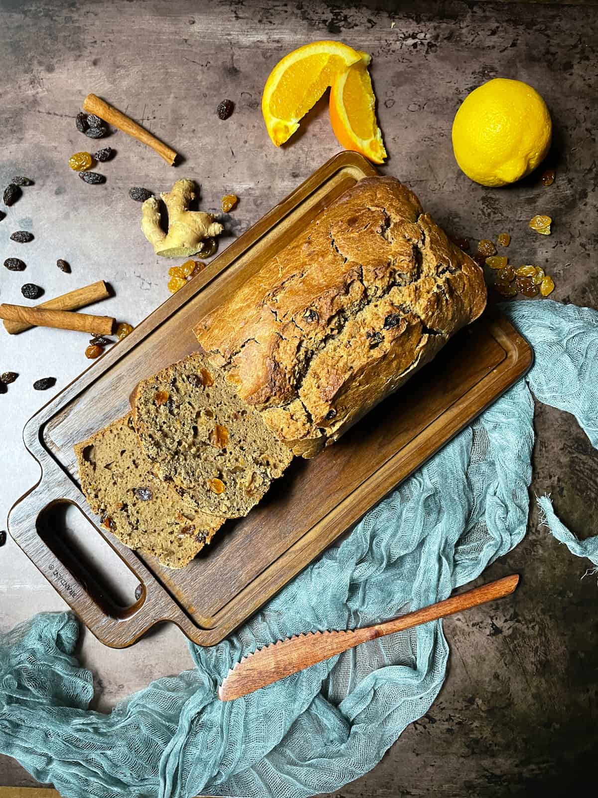 Dried fruit bread on a wood cutting board with a blue cloth, cinnamon, ginger, and raisins.