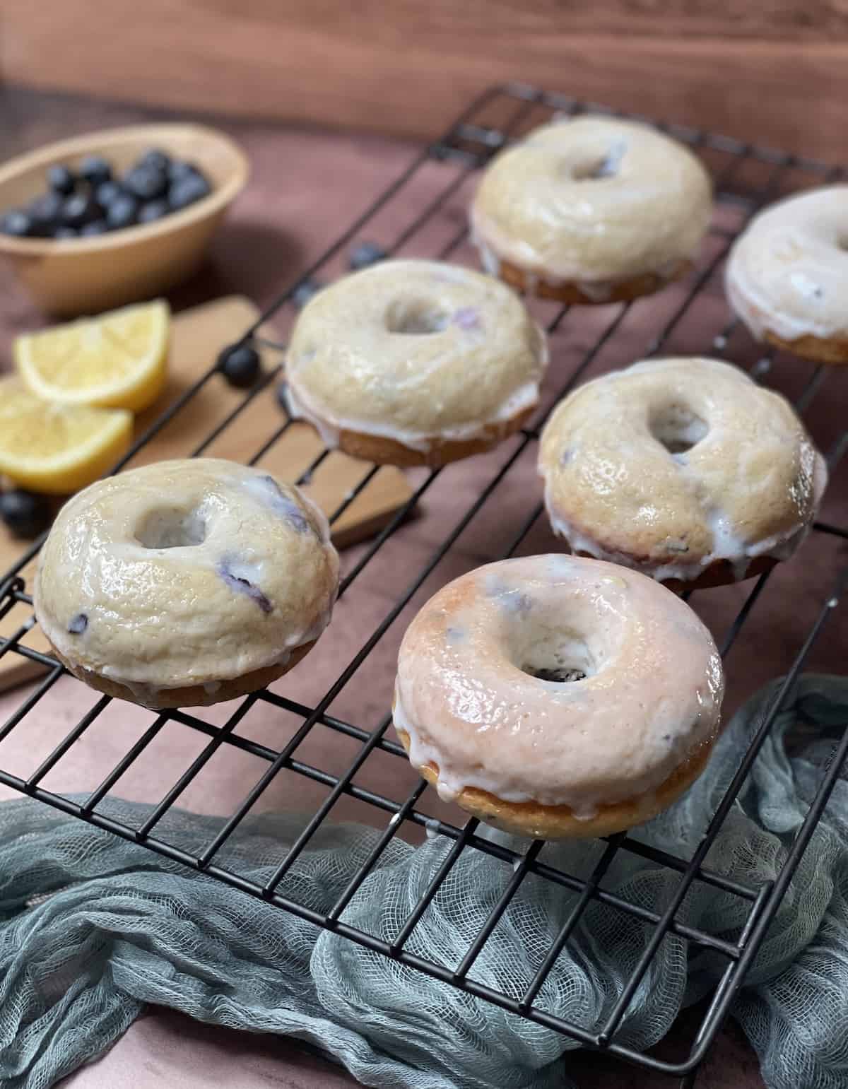 Iced baked blueberry cake donuts on a wire rack with lemons and blueberries on the side.