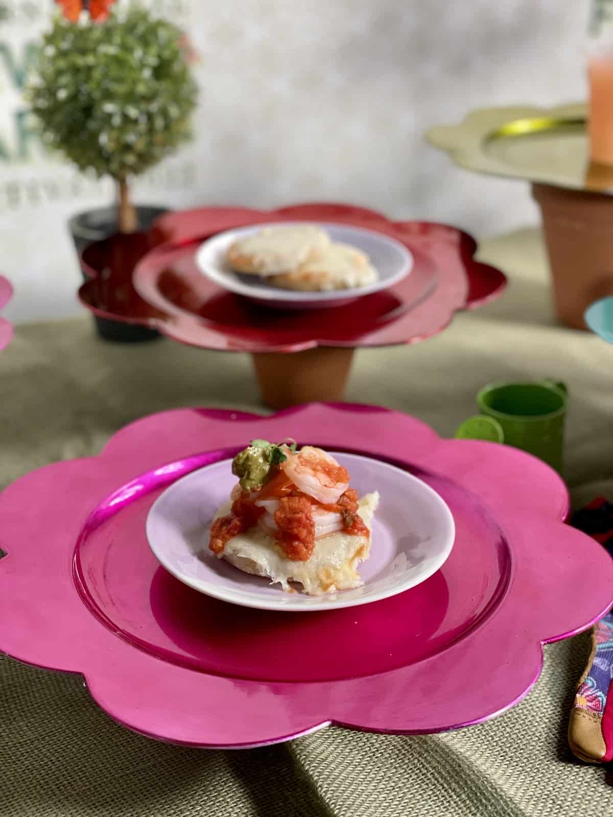 Arepa topped with Shrimp, crushed avocado aji (light pepper sauce), and tomato sauce on a pink plate.