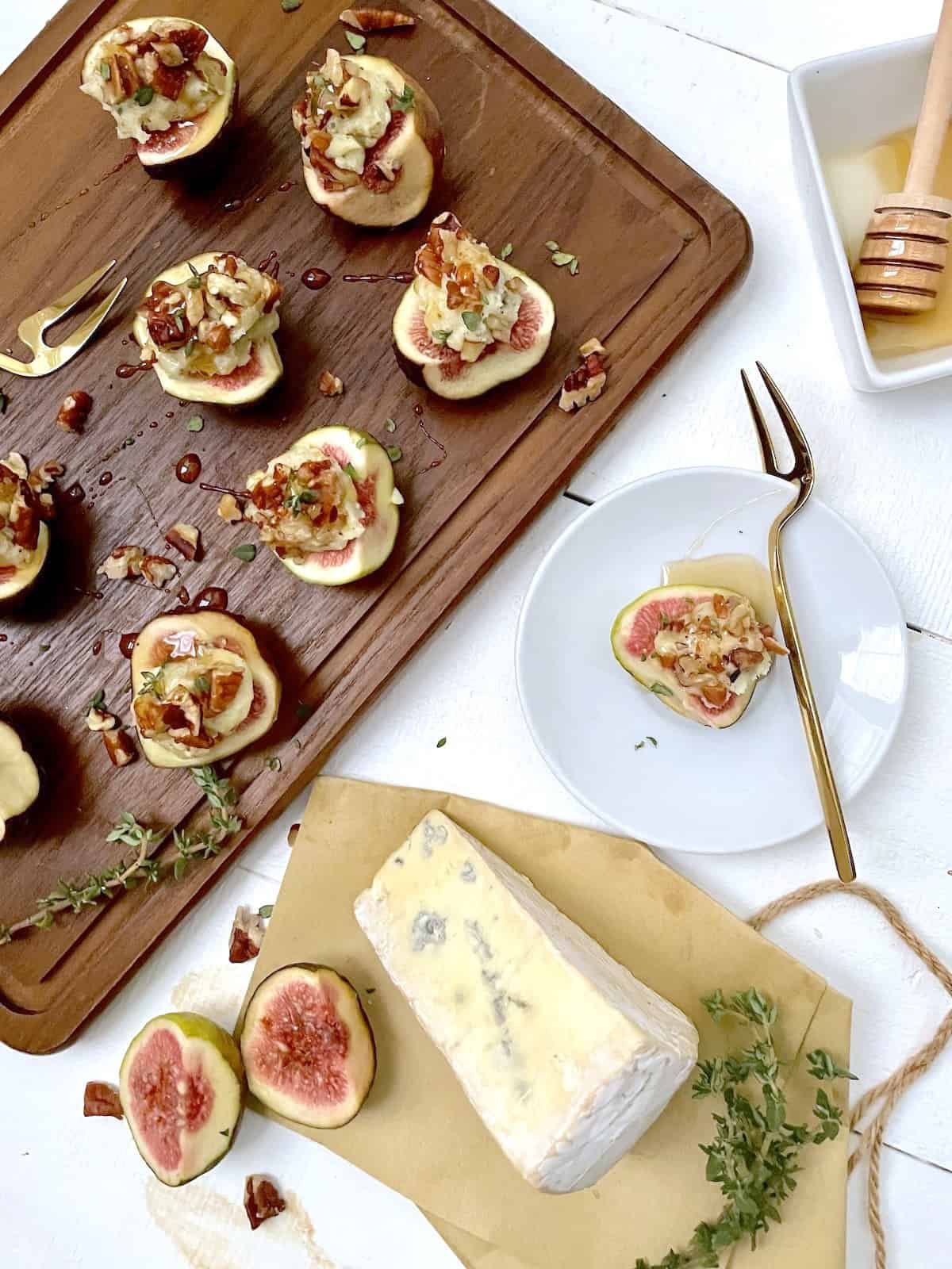 Figs with blue cheese, rosemary, nuts, and honey on a cutting board with block of cheese on paper.