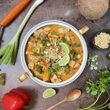 Thai chicken curry in a white bowl topped with peanuts, cilantro, and scallions and surrounded by fresh vegetables.