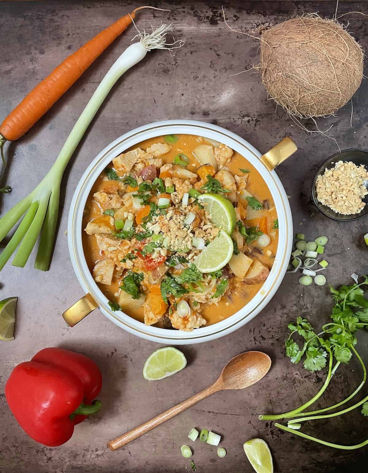 Thai chicken curry in a white bowl with red pepper, cilantro, onion, limes, peanuts, carrots, and a wooden spoon on a slate table.