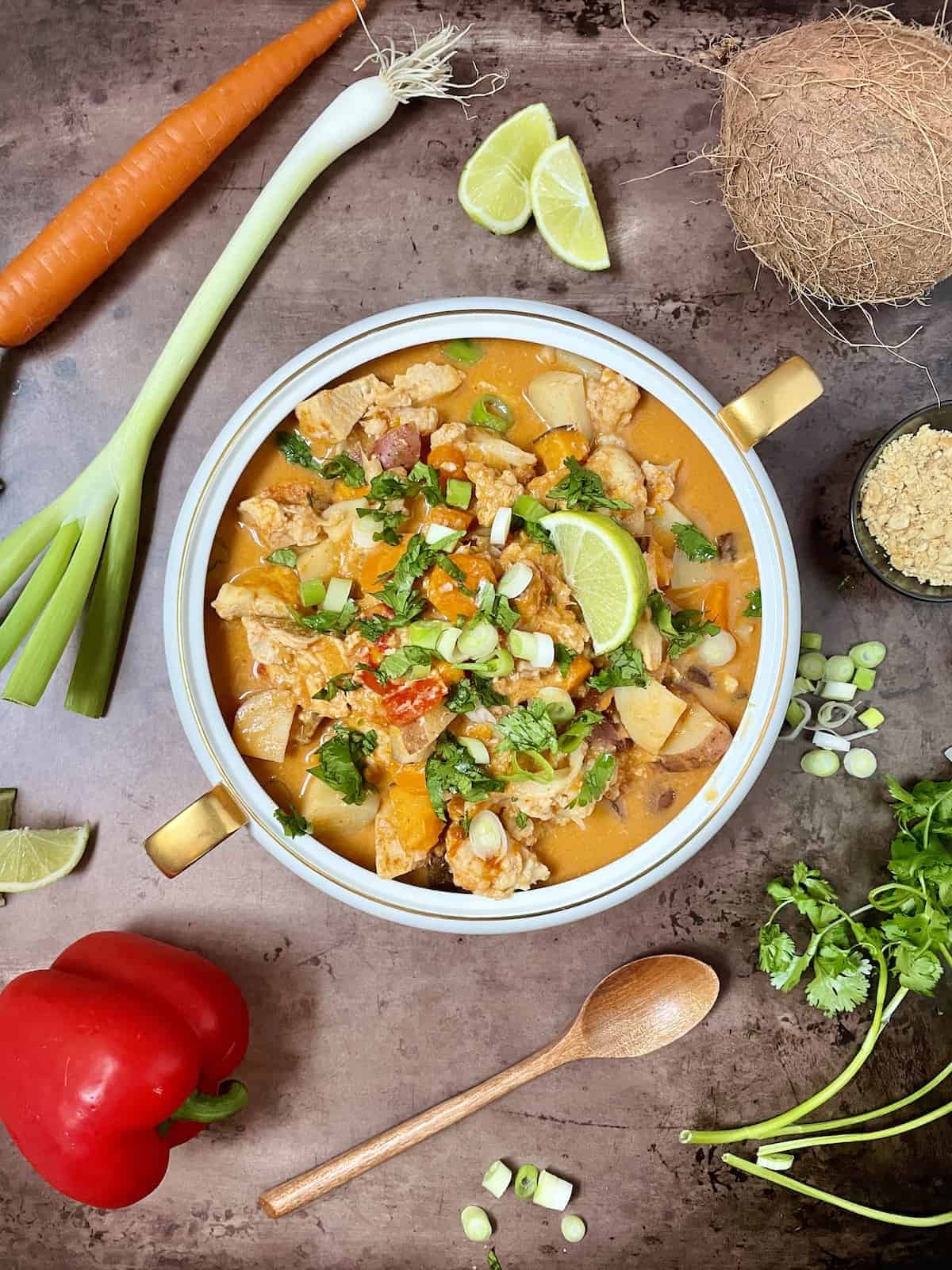 Thai chicken curry in a white bowl with fresh vegetables all around with a wooden spoon on slate table.
