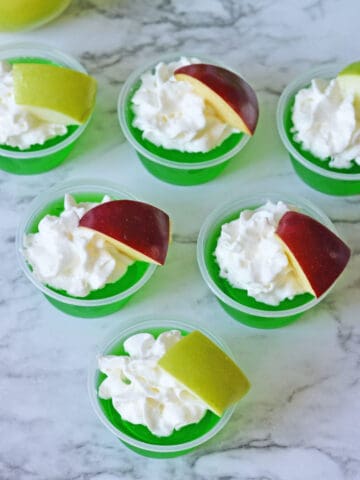 Green jello shots in plastic cups with whipped cream and apple slices.