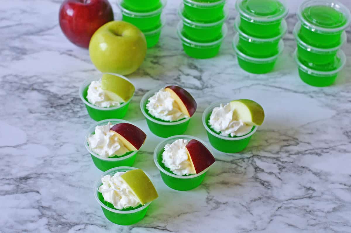 Green jello in plastic cups with whipped cream and apple slices.