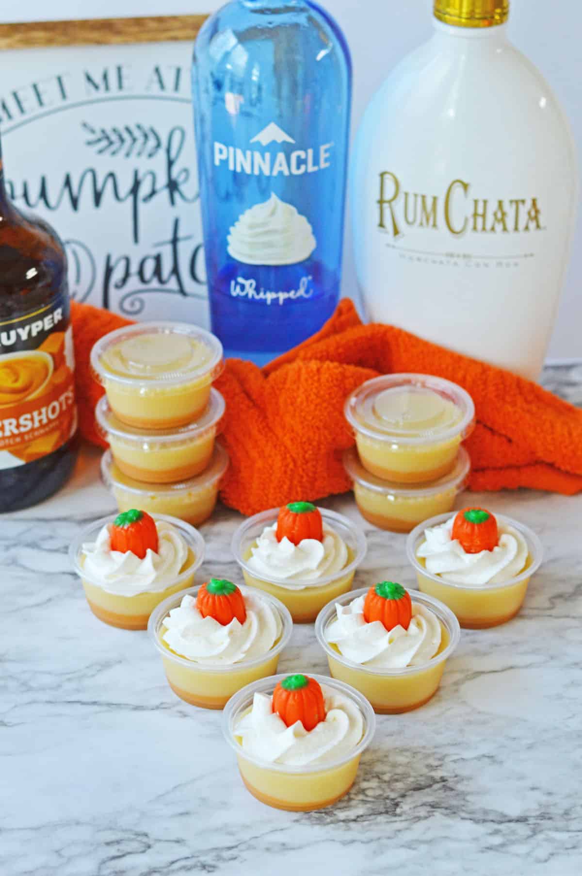 Pumpkin jello shots with whipped cream topped with pumpkin candy, with vodka, rum, and schnapps in background.