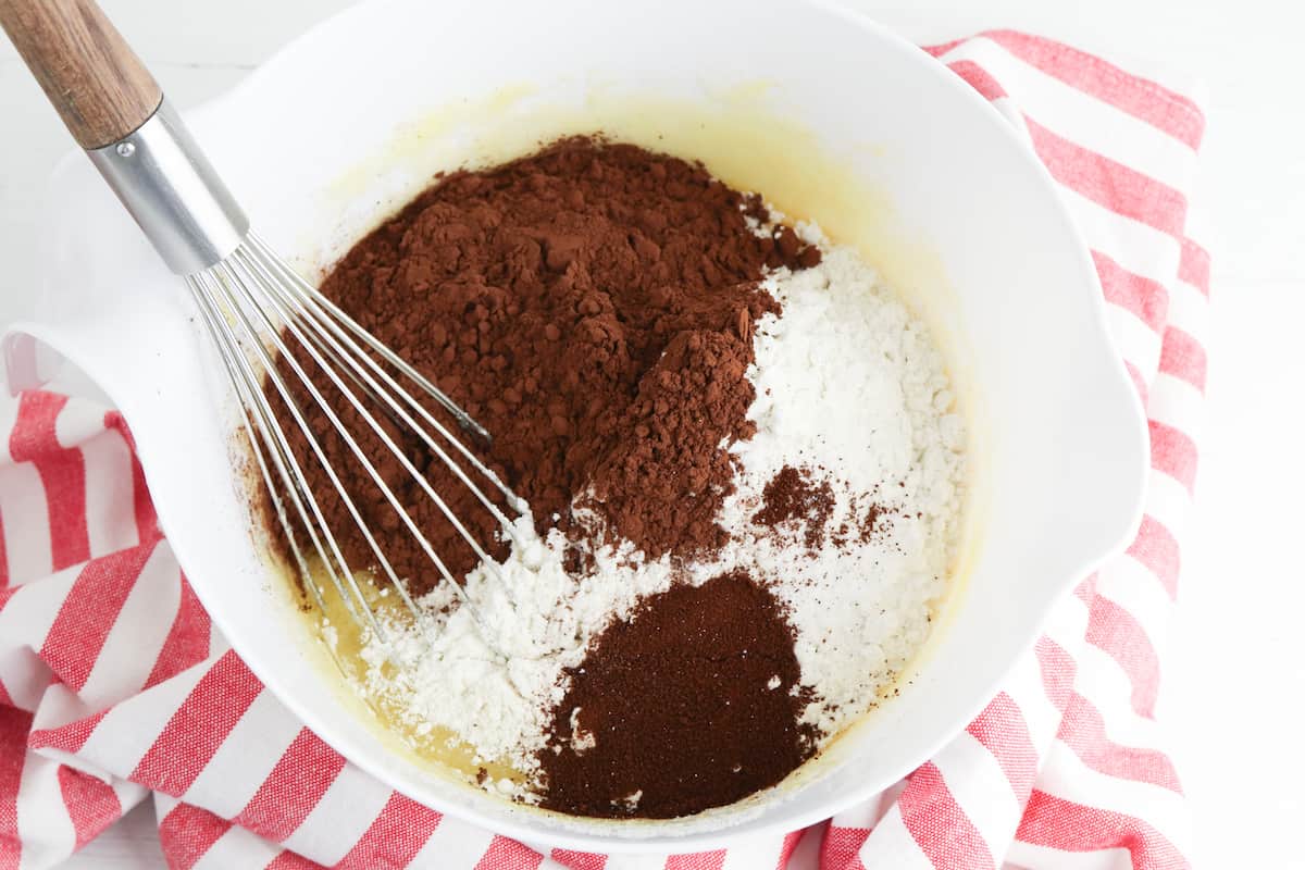 Spices and cocoa powder added to egg mixture in white bowl with whisk.