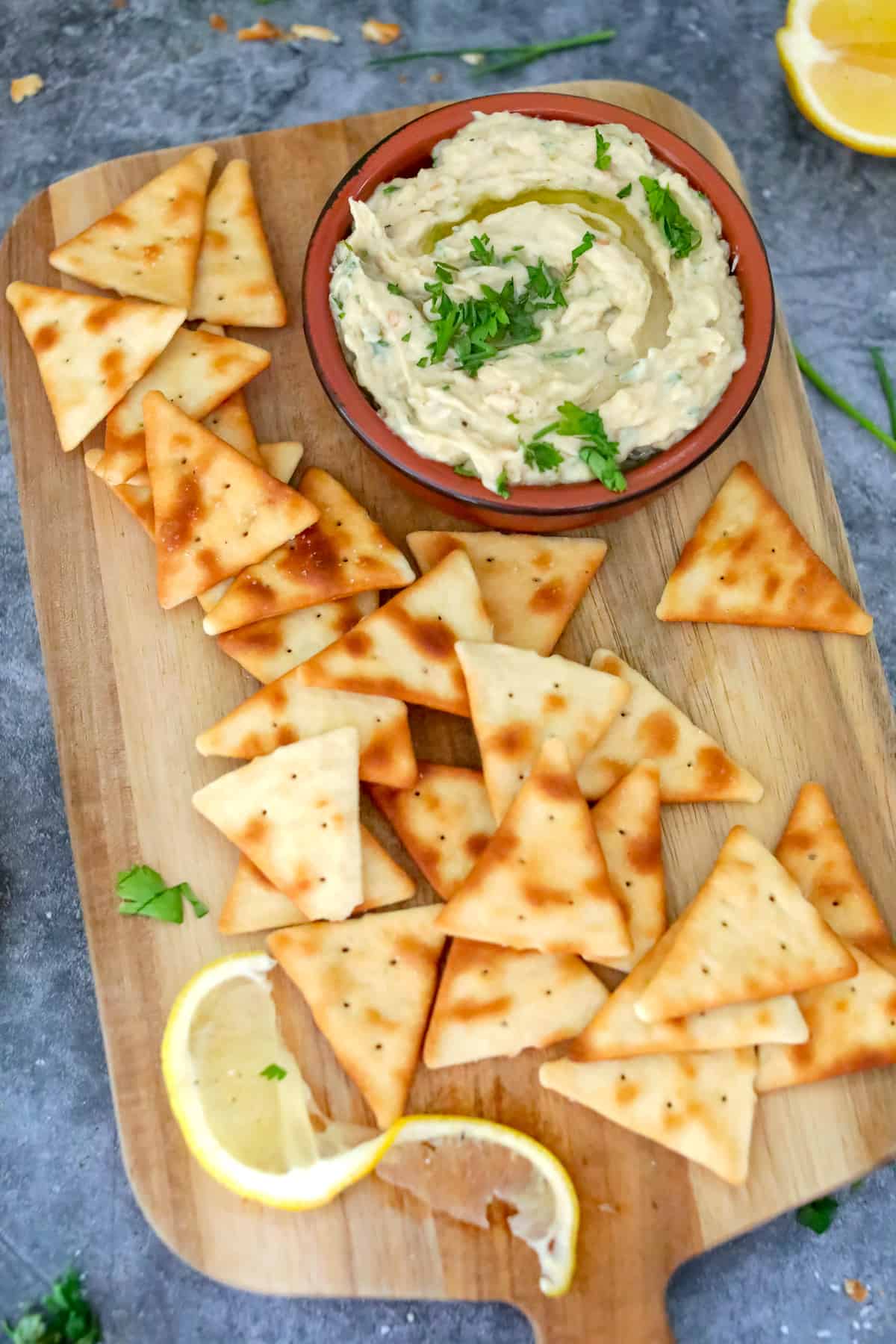White dip in a bowl on a wood cutting board with pita chips and lemon.