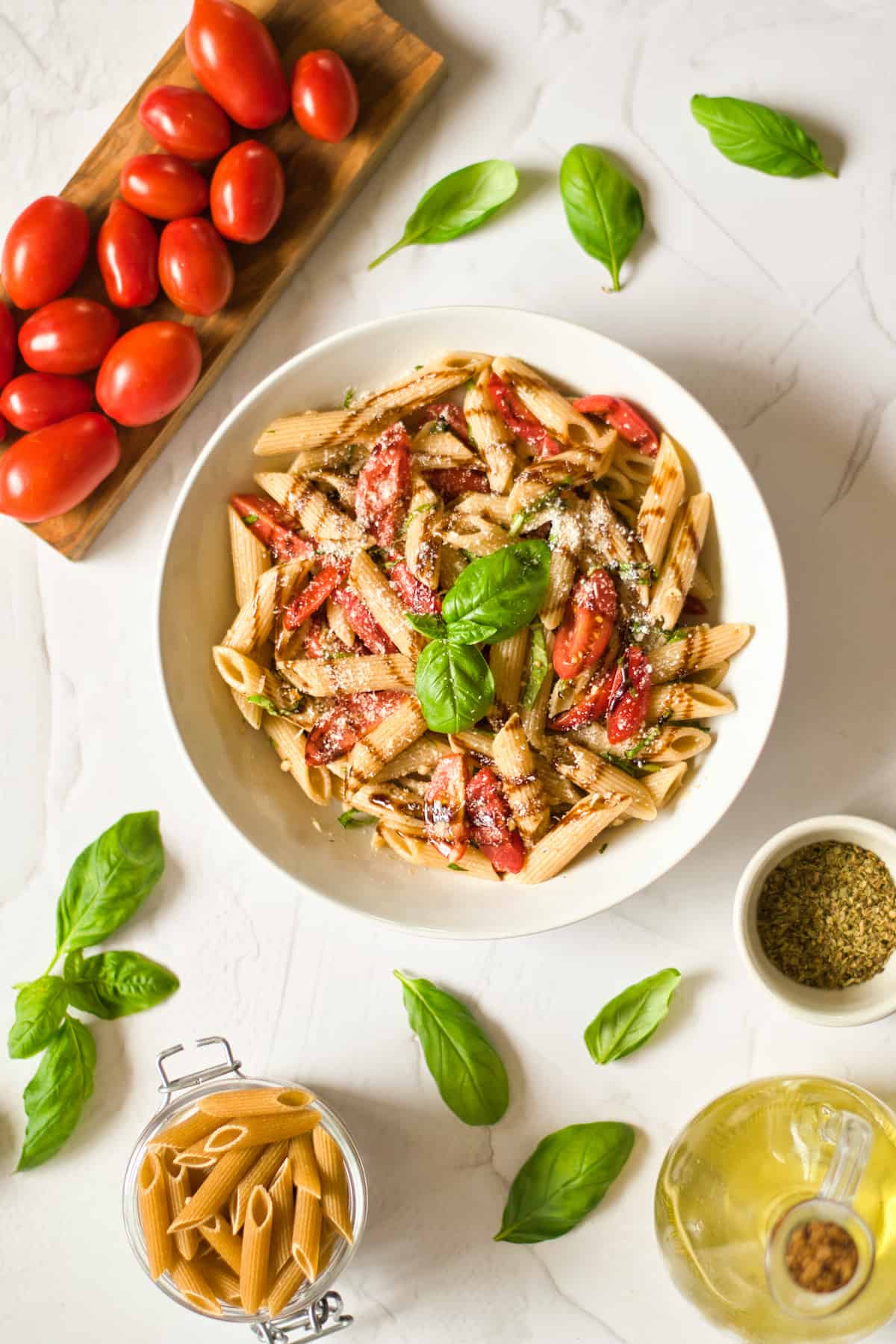 Pasta with tomatoes and basil in a white bowl on dinner table with bottle of oil, jar of pasta, basil leaves, seasoning, and tomatoes.