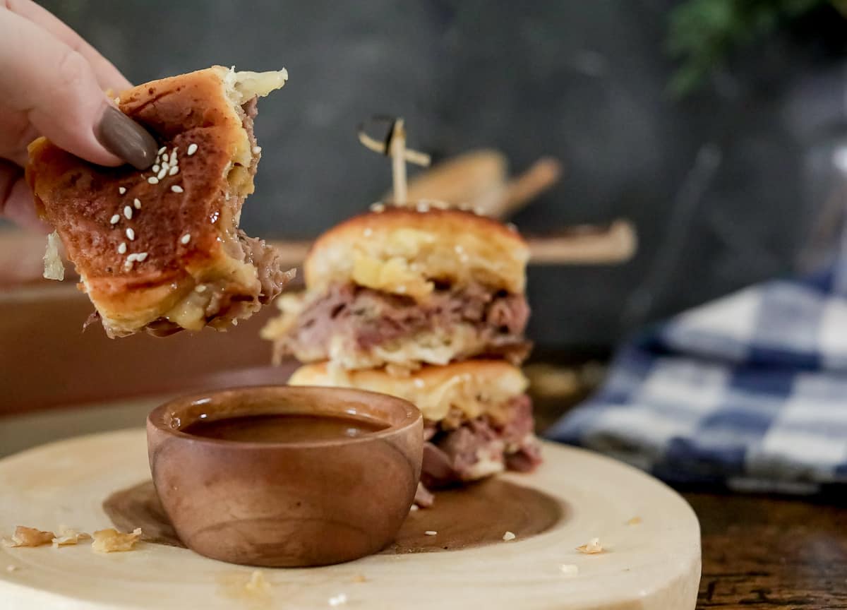 Two small roast beef sliders are stacked on a wooden board, secured with a toothpick. Another slider is in the background. The sliders have sesame seed buns and appear to be served in a rustic setting.