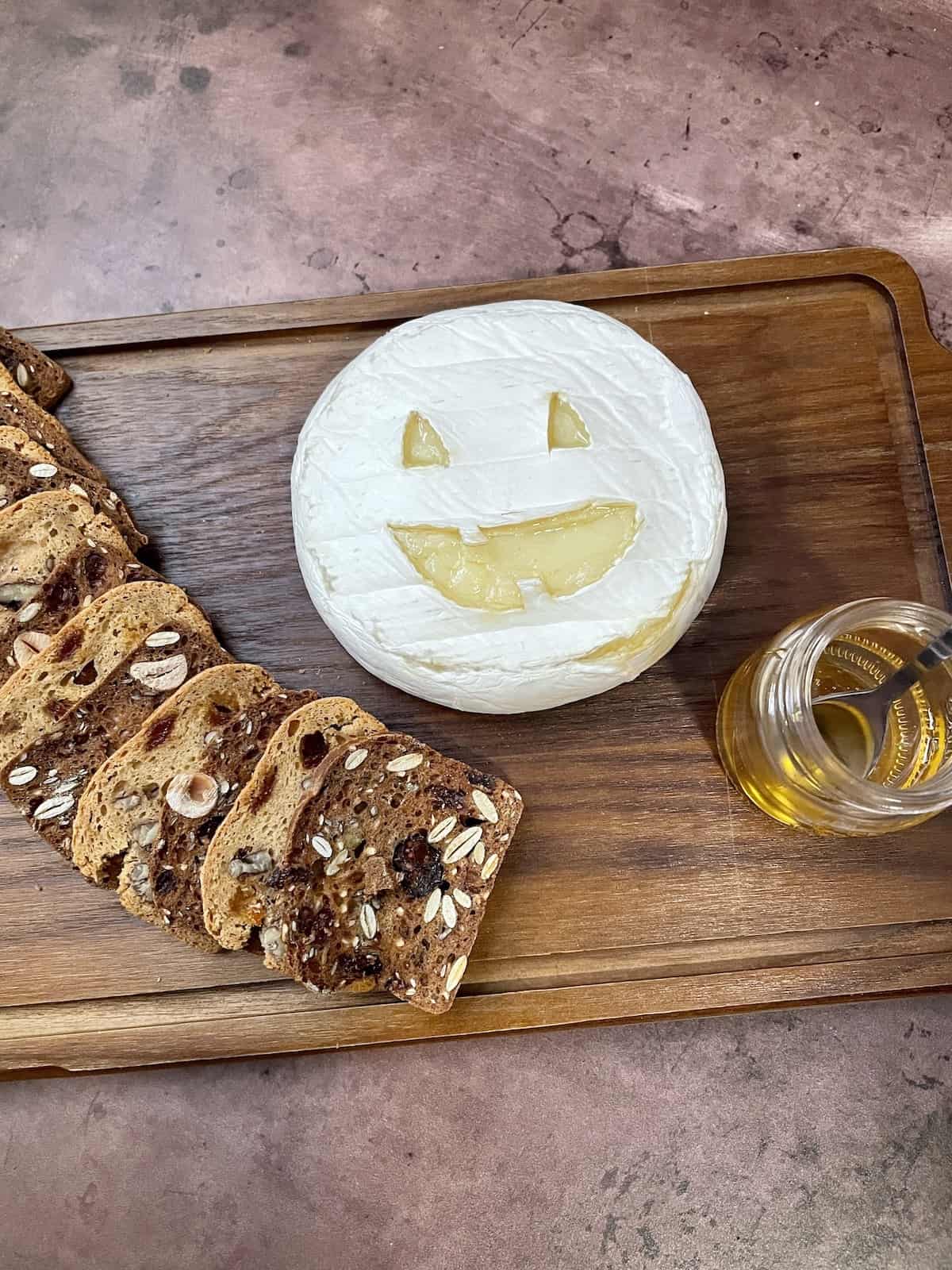 Brie cheese carved like a Jack O'Lantern on a wood board with crackers and honey.