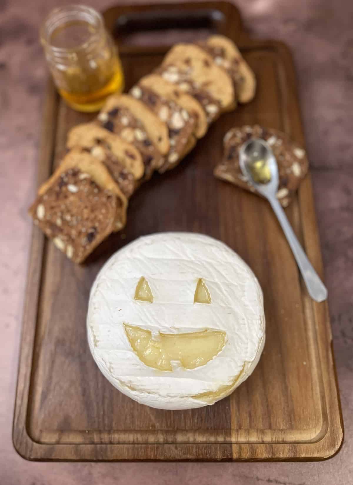 Brie cheese carved like a Jack O'Lantern on a wood board with crackers and honey.