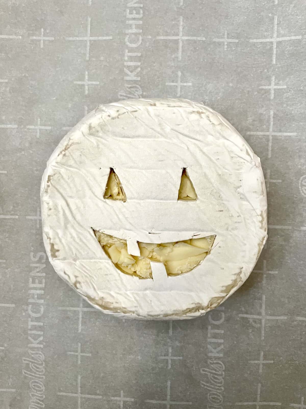 Brie with Jack O'Lantern face carved out on parchment paper on baking sheet.