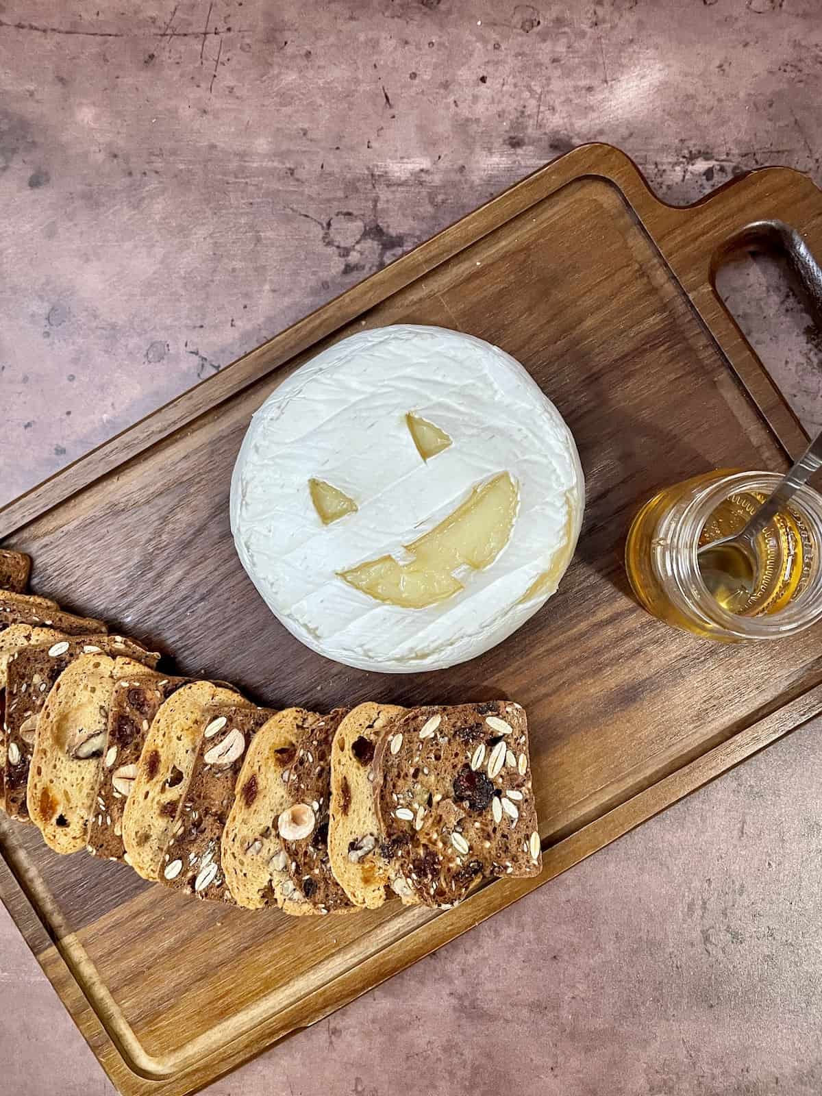 Brie cheese carved like a Jack O'Lantern on a wood board with crackers and honey.
